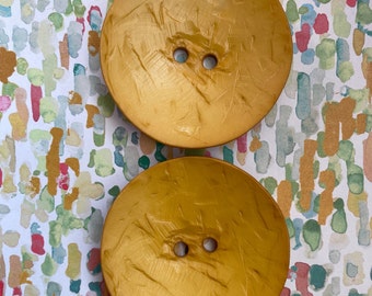 May include: Two large, round, gold-colored buttons with a textured surface. The buttons have two holes each.