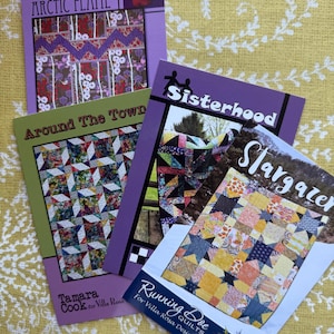 May include: A collection of quilt pattern books with titles including "Arctic Flame," "Around The Town," "Sisterhood," and "Stargazer." The covers feature colorful quilt designs and text. The books are on a yellow floral patterned surface.