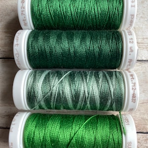 May include: Four spools of green thread, two solid green, one green and white ombre, and one solid dark green. The spools are labeled with the brand name "Sulky" and the article number "Art. 712-0051", "Art. 712-4037", and "1051".