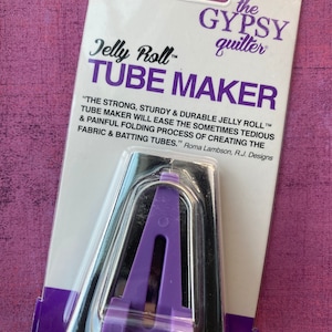 May include: Purple plastic tube maker tool for quilting, labeled "Jelly Roll™ TUBE MAKER" and "The Gypsy Quilter". The text reads "The strong, sturdy & durable Jelly Roll™ Tube Maker will ease the sometimes tedious & painful folding process of creating the fabric & batting tubes." Roma Lambson, R.J. Designs