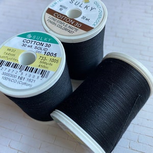 May include: Two spools of black Sulky Cotton 30 thread. The spools are labeled with the brand name, thread type, weight, color number, and yardage. The spools are on a white brick background.