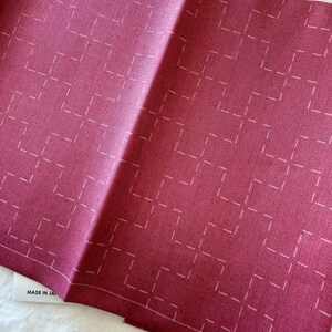 May include: A folded piece of burgundy fabric with a white geometric pattern. The fabric has a subtle texture and the pattern is made up of small squares with a white line in the center of each square. The fabric has a label that says "MADE IN JAPAN".