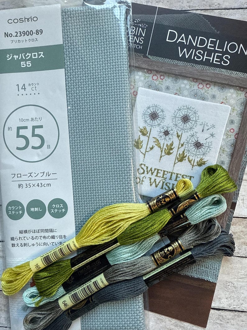 May include: A cross-stitch kit with a light blue fabric, embroidery floss in various shades of green and gray, and a pattern that says "Dandelion Wishes". The kit includes aida cloth, floss, and instructions for a counted cross-stitch project.