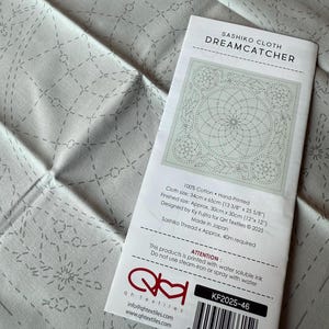 May include: A white Sashiko cloth with a printed design of a dreamcatcher. The cloth is 13 3/8 inches by 25 5/8 inches and the finished size is 12 inches by 12 inches. The cloth is designed by Ky Fujita for QH Textiles and is made in Japan. The product is printed with water soluble ink and should not be steam ironed or sprayed with water. The product number is KF2025-46.