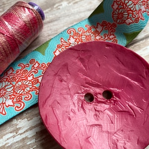 May include: A large, round, pink button with two holes in the center. The button has a textured surface and a shiny finish.