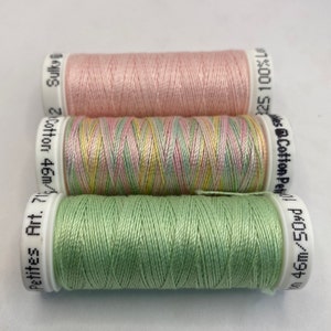 May include: Three spools of thread, one pink, one green, and one multi-colored with pink, green, and yellow. The spools are labeled "Cotton Pet 25 100%", "Petites Art. 46m/2", and "Wes @Cotton Pet PROS/W96W".