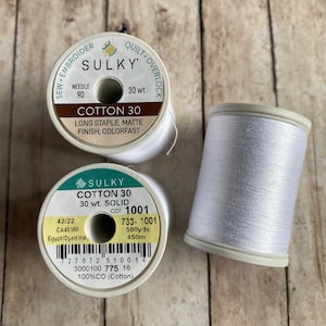 May include: Two spools of white cotton thread, one labeled "Sulky Cotton 30 Long Staple, Matte Finish, Colorfast" and the other labeled "Sulky Cotton 30 Solid 30 wt. col: 1001 733-1001 500yds 450m 100%CO (Cotton)"
