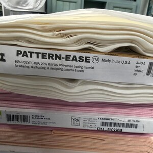 Op de afbeelding: Een stapel van drie rollen stof. De bovenste rol is wit Pattern-Ease niet-geweven overtrekpapier van 117 cm breed. De middelste rol is roze Essex Blossom stof van 114 cm breed. De onderste rol is een effen licht perzikkleurige stof.
