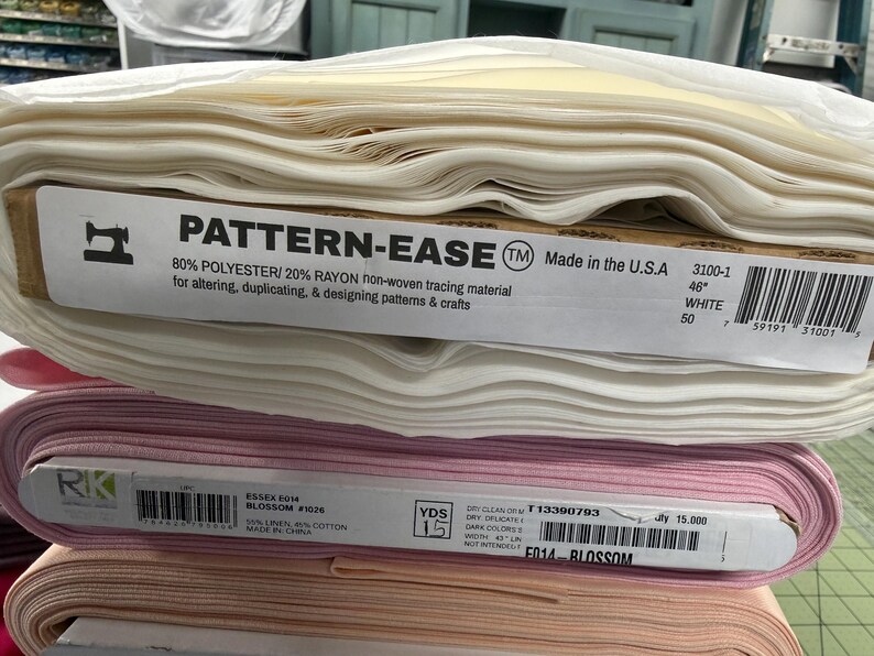 Op de afbeelding: Een stapel van drie rollen stof. De bovenste rol is wit Pattern-Ease niet-geweven overtrekpapier van 117 cm breed. De middelste rol is roze Essex Blossom stof van 114 cm breed. De onderste rol is een effen licht perzikkleurige stof.