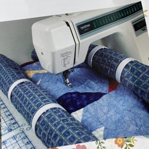 May include: A white Brother sewing machine with a blue and white quilt being sewn. The quilt is being fed through two blue and white fabric rollers with white plastic bands.