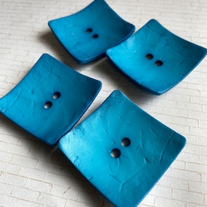 May include: Four square-shaped buttons with a textured surface. The buttons are a vibrant teal blue color and have two holes each.