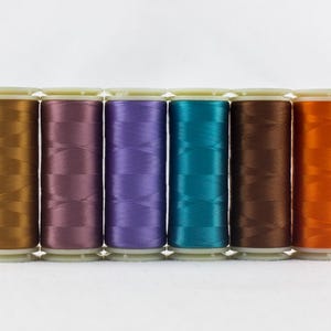 May include: A row of six spools of thread in various colors: gold, purple, teal, brown, and orange. The spools are cylindrical with white plastic bases and tops, and the thread is tightly wound.