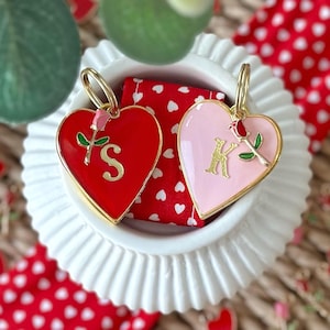 May include: Two heart-shaped pet tags with gold rings. The red tag has a gold letter "S" and a gold rose with green leaves. The pink tag has a gold letter "K" and a gold rose with green leaves.