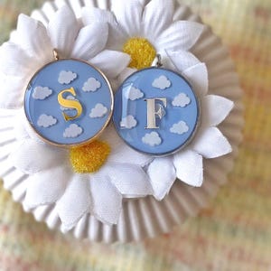 May include: Two circular pendants featuring a sky blue backdrop with white cloud details. One pendant has a gold letter "S", the other a silver "F". Both are set in a metal frame.
