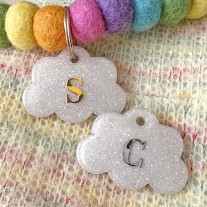 May include: Two cloud-shaped pet tags with glittery white surfaces. One tag has a gold letter "S", the other a silver letter "C". A rainbow-coloured felt ball collar is in the background.