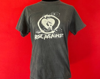 May include: Black t-shirt with a white graphic of a fist in a heart with a circle around it. The text "Rise Against" is below the graphic.