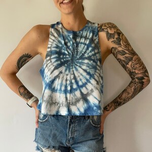 May include: A blue and white tie-dye cropped tank top with a spiral pattern. The tank top is worn with denim shorts.