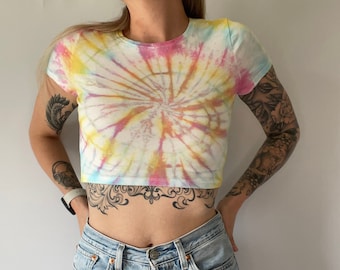 May include: A white crop top with a tie-dye pattern in shades of pink, yellow, and blue. The shirt has short sleeves and a round neckline.