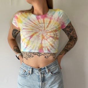 May include: A white crop top with a tie-dye pattern in shades of pink, yellow, and blue. The shirt has short sleeves and a round neckline.