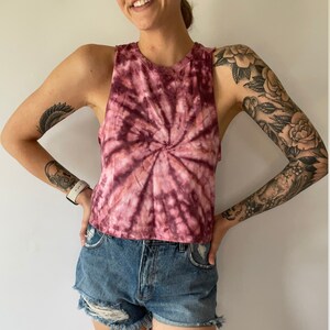 May include: A cropped tie-dye tank top in shades of pink and burgundy with a spiral pattern. The tank top is sleeveless and has a relaxed fit.