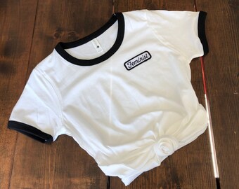 May include: White t-shirt with black trim around the neckline and sleeves. The shirt has a black patch with the word "Feminist" on it.