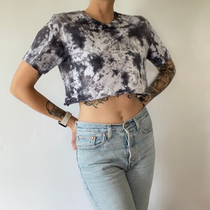 May include: A black and white tie-dye cropped t-shirt with a relaxed fit. The shirt has a raw edge hem and is made of a soft, comfortable fabric.