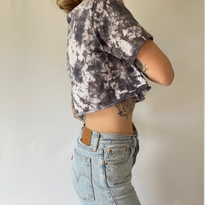 May include: A woman wearing a cropped tie-dye shirt in shades of black and white with a pair of light blue denim jeans.