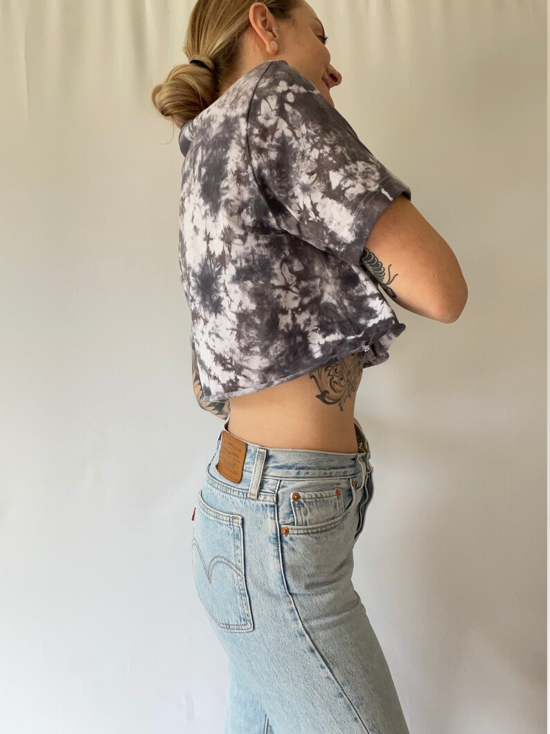 May include: A woman wearing a cropped tie-dye shirt in shades of black and white with a pair of light blue denim jeans.
