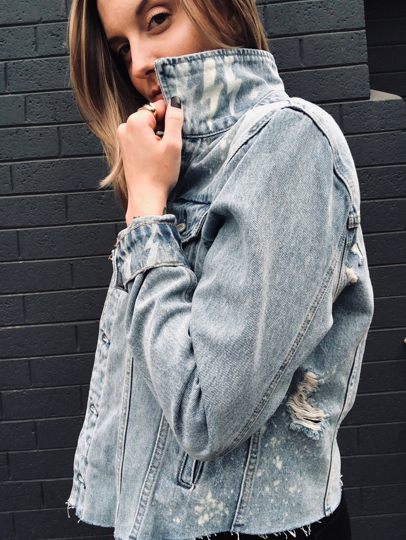 May include: A light blue denim jacket with white paint splatters and distressed details. The jacket has a high collar and a button closure.