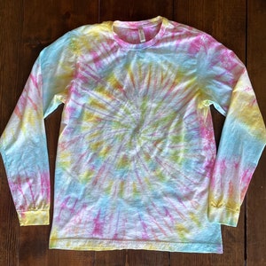 May include: A long-sleeved shirt with a tie-dye pattern in shades of pink, yellow, and blue. The shirt has a circular pattern with a swirl design.