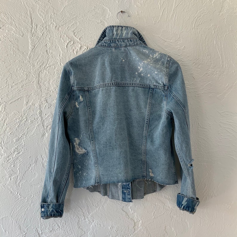 May include: A light blue denim jacket with distressed details and white paint splatters. The jacket has a classic collar, button closure, and long sleeves.