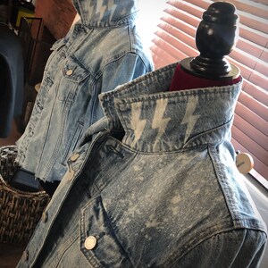 May include: Two denim jackets with a light blue wash and white lightning bolt designs on the collars. The jackets are displayed on mannequins.