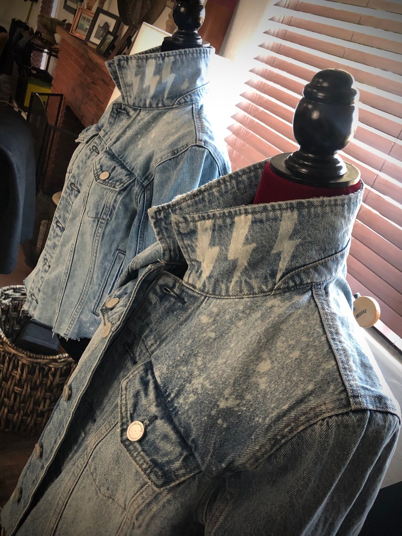 May include: Two denim jackets with a light blue wash and white lightning bolt designs on the collars. The jackets are displayed on mannequins.