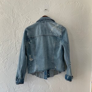 May include: A light blue denim jacket with white paint splatters and distressed details. The jacket has a button-up front and long sleeves.