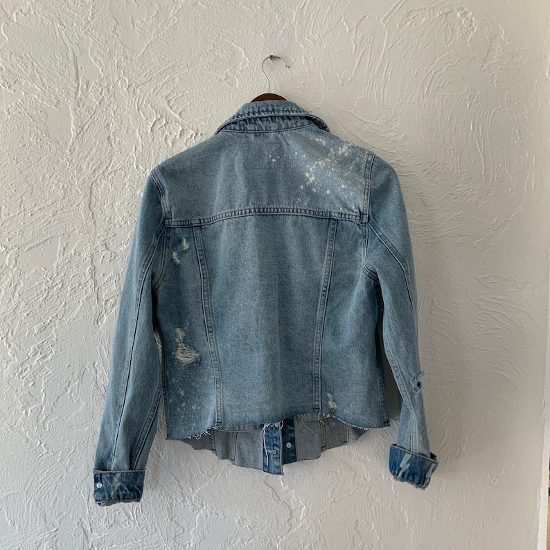 May include: A light blue denim jacket with white paint splatters and distressed details. The jacket has a button-up front and long sleeves.
