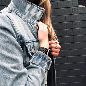 May include: A light blue denim jacket with a white lightning bolt design on the collar. The jacket is unbuttoned and the sleeves are rolled up. The person is wearing a black watch on their wrist.