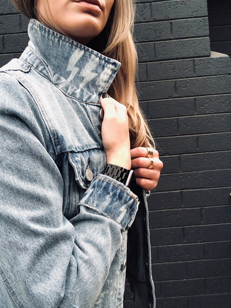 May include: A light blue denim jacket with a white lightning bolt design on the collar. The jacket is unbuttoned and the sleeves are rolled up. The person is wearing a black watch on their wrist.