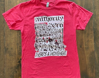May include: Red t-shirt with a black and white graphic print of a crowd of people with red heads. The text "authority zero" and "there's a movement" is printed on the shirt.