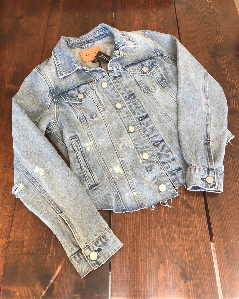 May include: Light blue denim jacket with distressed details, including a frayed hem and a ripped sleeve. The jacket has a button closure and multiple pockets.