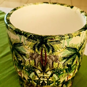 Puede incluir: Un macetero de cerámica blanca con un estampado de jungla verde, amarillo y marrón con palmeras y elefantes.