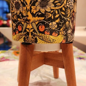 May include: A ceramic planter with a floral and bird design in shades of yellow, black, and green. The planter is on a wooden stand with three legs.