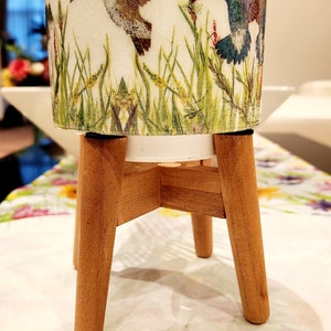 Könnte beinhalten: Ein weißer Keramiktopf mit einem Kolibri-Design. Der Topf steht auf einem Holzständer mit drei Beinen.
