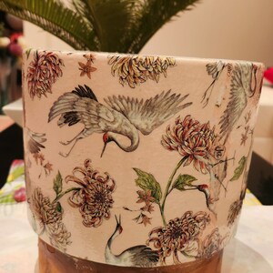 May include: A ceramic planter with a pink background and a floral design featuring cranes, flowers, and leaves. The planter has a wooden base.