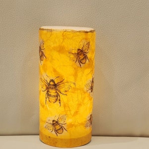 May include: A yellow cylindrical candle holder with a textured surface and a pattern of black bees. The candle holder is lit and glowing.