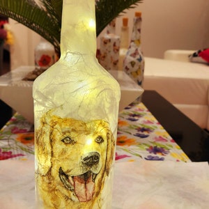 Può includere: Una bottiglia di vetro bianco con un'immagine di un cane golden retriever e luci a stringa all'interno. La bottiglia ha un tappo di sughero e si trova su una superficie con motivo floreale.