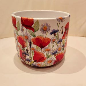 May include: White ceramic flower pot with a red, white and blue floral pattern. The pot is decorated with red poppies and other wildflowers.