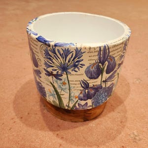 May include: A ceramic plant pot with a floral design featuring blue irises, agapanthus, and dragonflies. The pot has a white interior and a wooden base. The exterior is decorated with script and botanical illustrations.