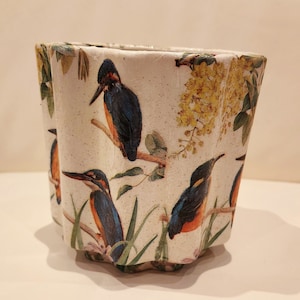 May include: A white ceramic planter with a scalloped rim features a colourful floral and bird design. The planter has a speckled white glaze and is decorated with a repeating pattern of blue, orange, and green birds perched on branches with yellow flowers.
