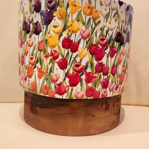 May include: A white ceramic planter with a floral pattern of colorful tulips. The planter has a brown wooden base.