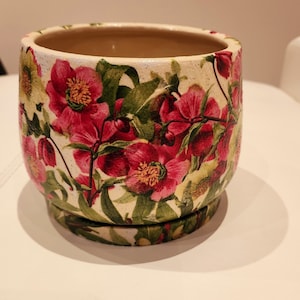 May include: A white ceramic flower pot with a floral design featuring pink and green flowers. The pot has a saucer.